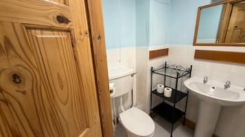 A bathroom with a wooden door, a toilet, a sink, and a shelf with toilet paper.
