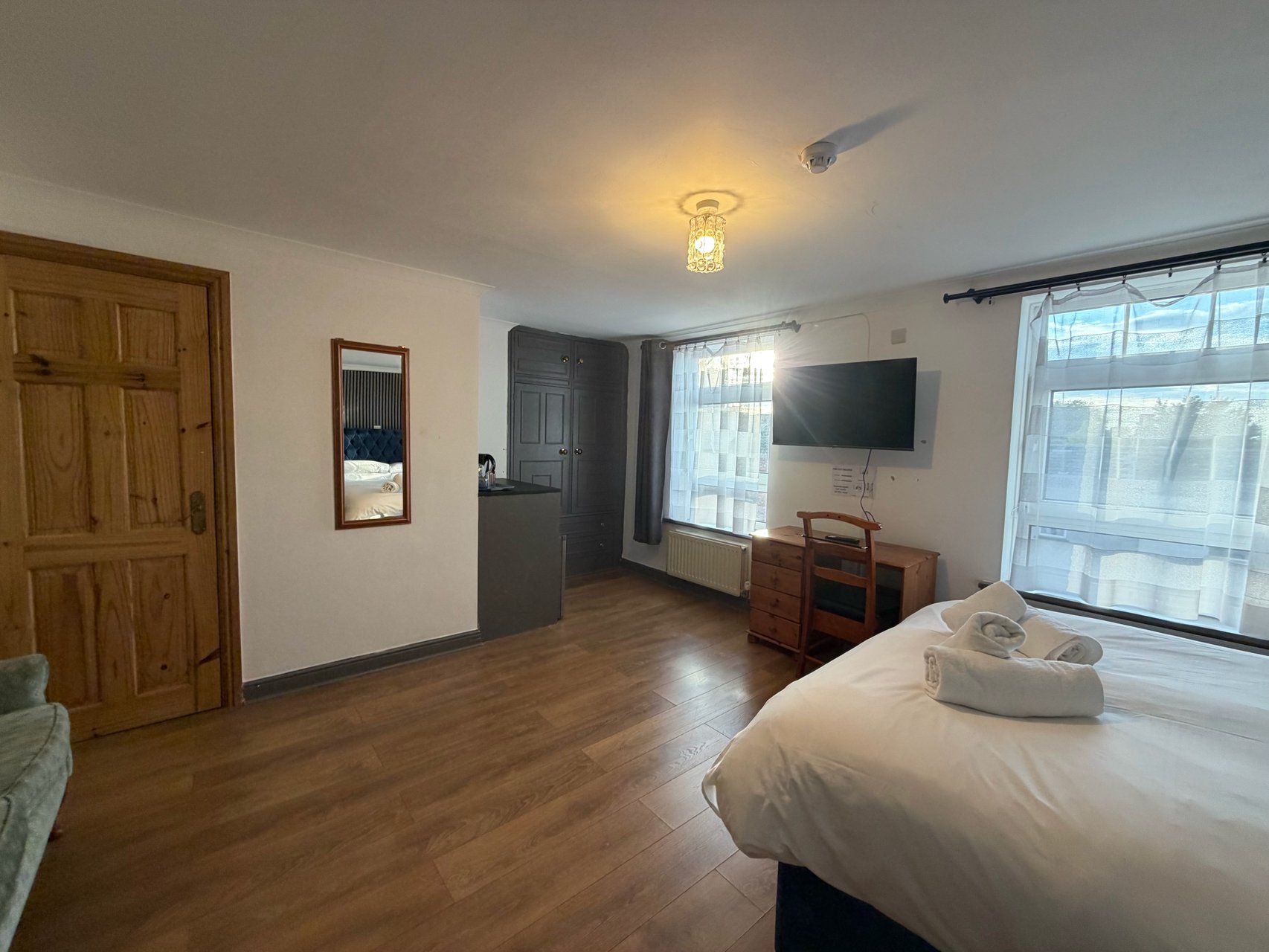 A hotel room with a wooden floor, a bed with a white duvet, a desk with a chair, a television mounted on the wall, and a window with curtains.