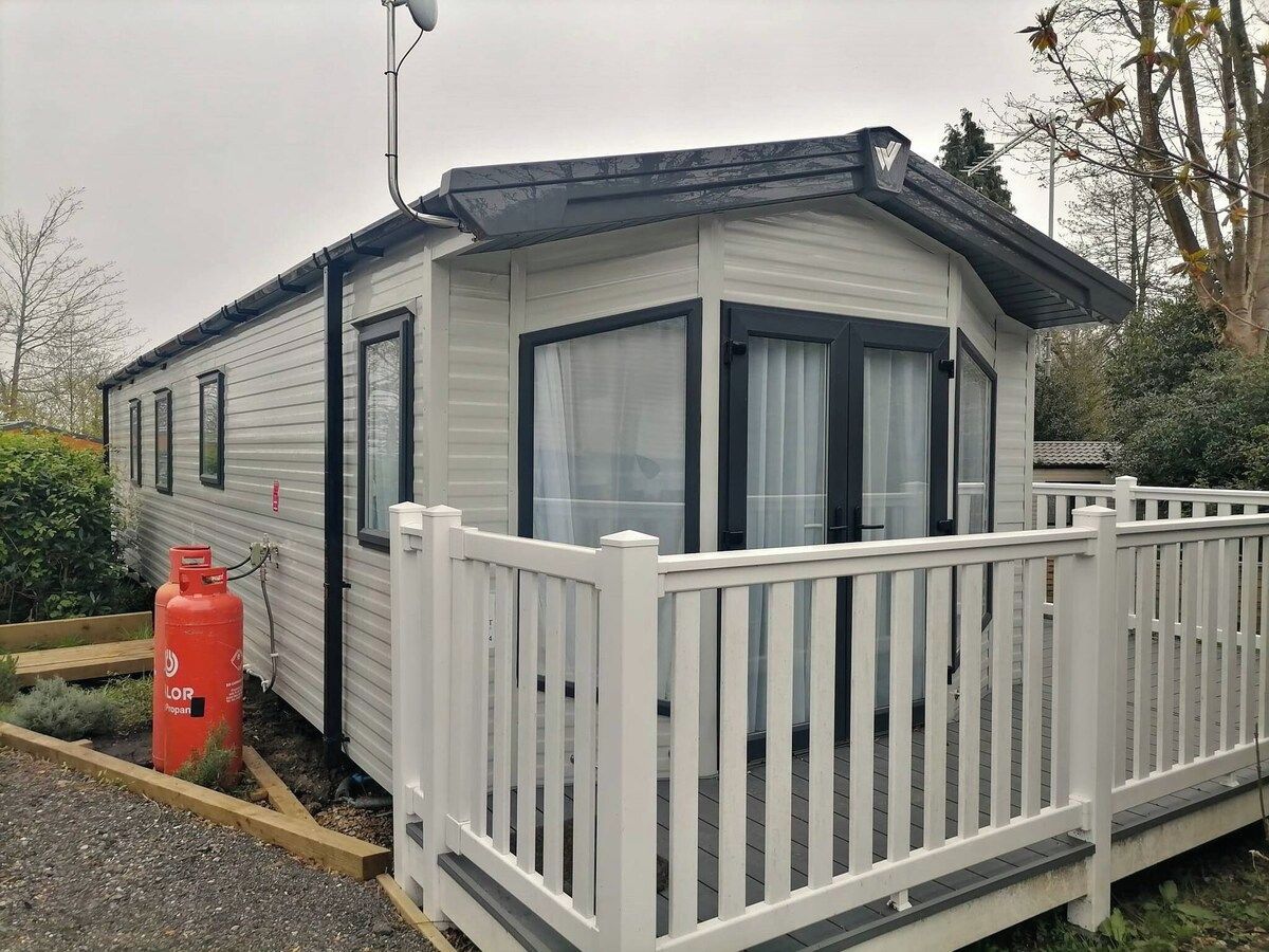 A small, single-story mobile home with a white exterior, a fenced porch, and a propane tank outside.