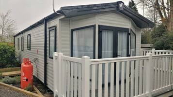 A small, single-story mobile home with a white exterior, a fenced porch, and a propane tank outside.