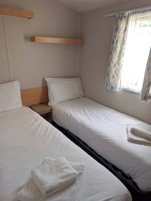 A single room with two beds, a wooden shelf, a window with a curtain, and a towel on one bed.