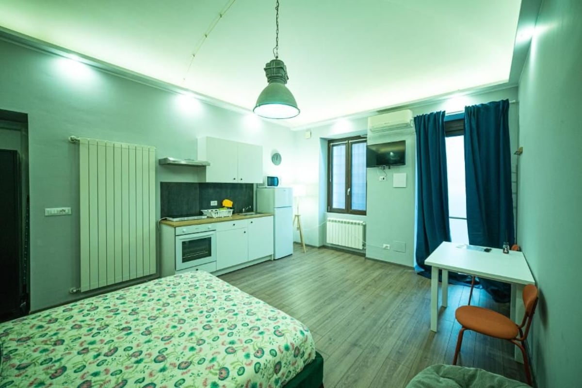 Une pièce de taille modeste comprenant un lit, un bureau, une chaise, un coin cuisine et une fenêtre avec des rideaux.