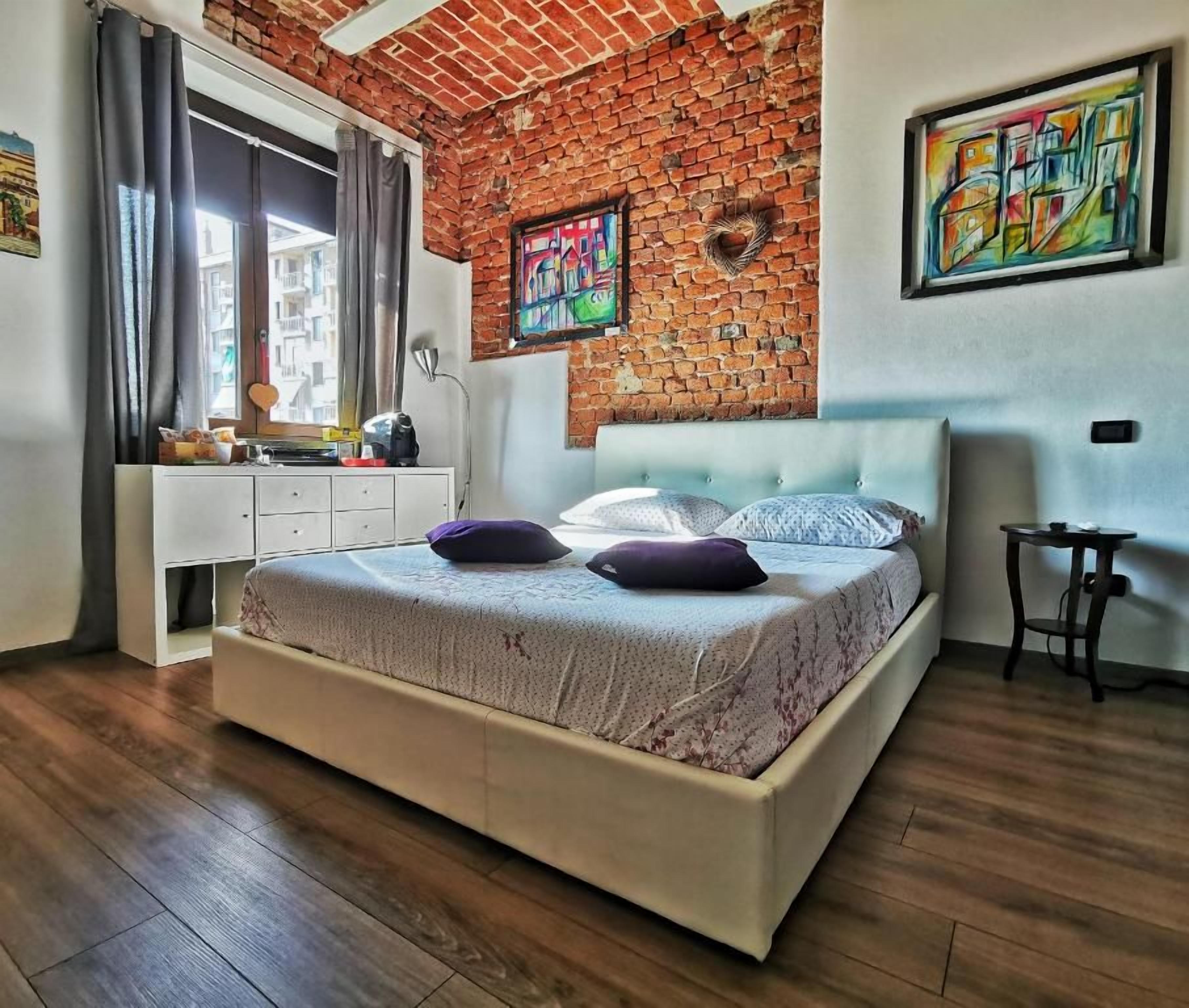 Une chambre avec un mur en briques, un lit recouvert d’une couvre-lit à motifs, une commode et des œuvres d’art accrochées aux murs.