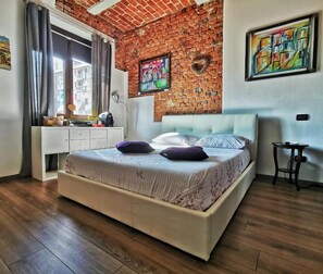 Une chambre avec un mur en briques, un lit recouvert d’une couvre-lit à motifs, une commode et des œuvres d’art accrochées aux murs.