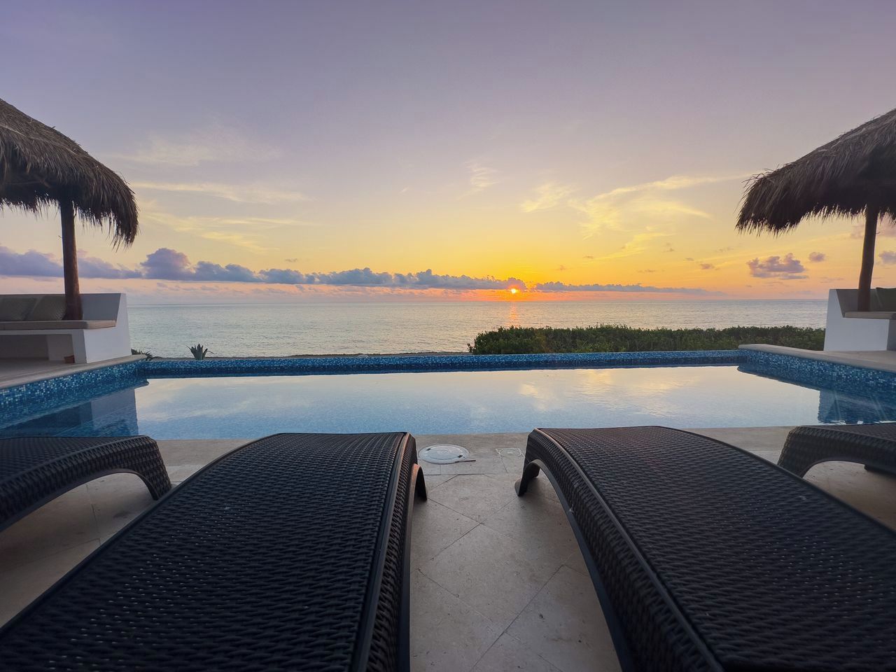 An infinity pool with lounge chairs and a view of the sunset over the ocean.