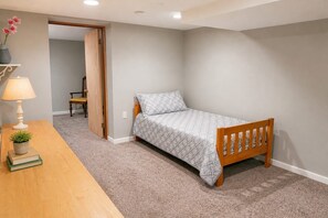 A bedroom with a single bed, a bedside table with a lamp, a small plant, and a chair in the background.