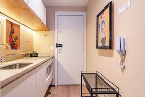 Een kitchenette met een spoelbak, fornuis en aanrecht. Er is een deur met slot en een klein tafeltje met een stoel. Een ingelijst beeld hangt aan de muur.