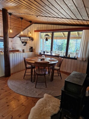 A cozy cabin interior with a wooden dining table, chairs, a fireplace, and a kitchen area with hanging lights and a window overlooking greenery. - New Listing (45751 Bullaren)