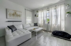 Ein modernes Wohnzimmer mit einem weißen Sofa, einem Couchtisch und einem schwarzen Teppich. Es gibt Fenster mit transparenten Vorhängen und eine Kristallleuchter.