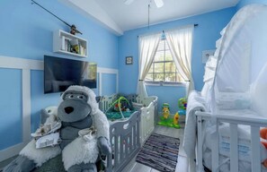 Ein Zimmer mit Babybett, Spielbereich mit Spielzeug, Fernseher und einem Fenster mit Vorhängen.