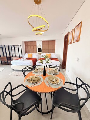 A modern hotel room with a dining area, two beds with orange pillows, a large window, and a hanging light fixture. - O Melhor de Goiania! Apt de 1 Quarto no coracao do Marista (Goiânia)