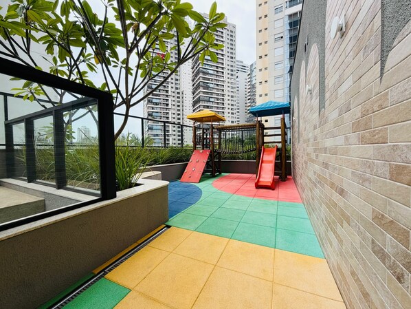 Ein Spielbereich für Kinder mit Rutschen und einer Kletteranlage, umgeben von einem Gebäude mit Backsteingemäuer und einem Balkon mit Fenstern.