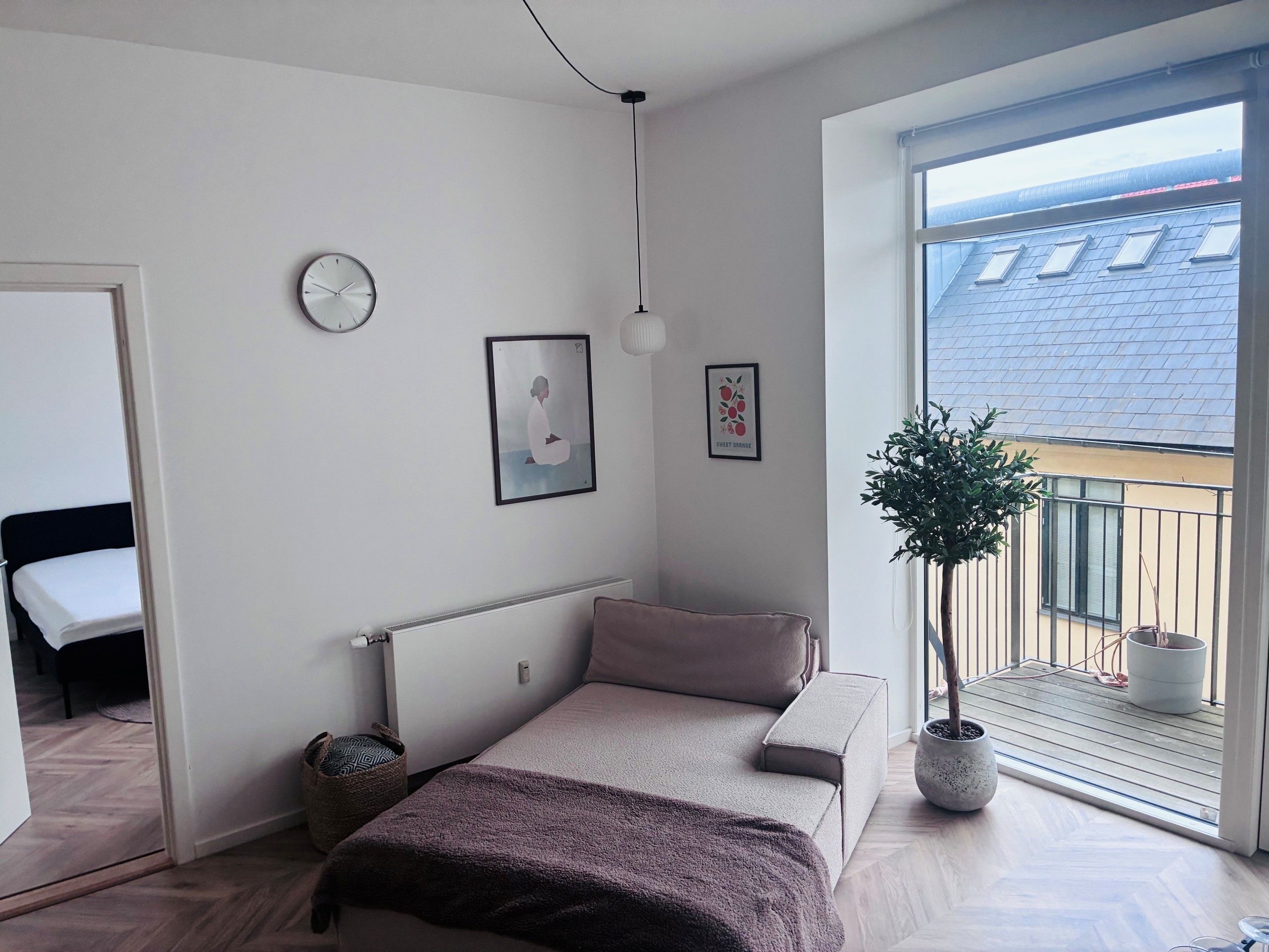 A bedroom with a bed, a bedside table, a wall clock, and a window with a balcony.