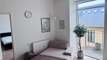 A bedroom with a bed, a bedside table, a wall clock, and a window with a balcony.