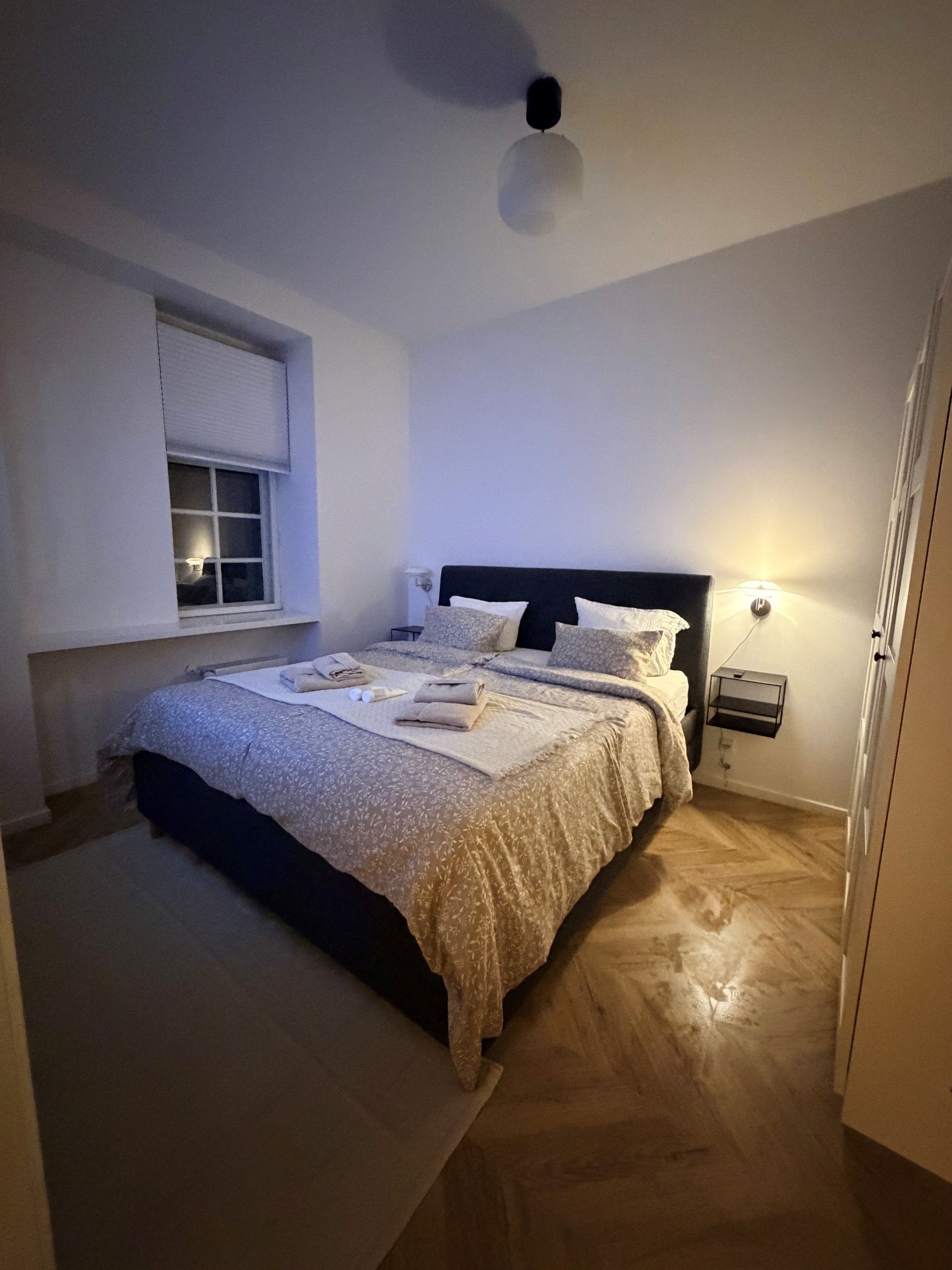 A bedroom with a bed, bedside tables, a window with blinds, and a ceiling light.