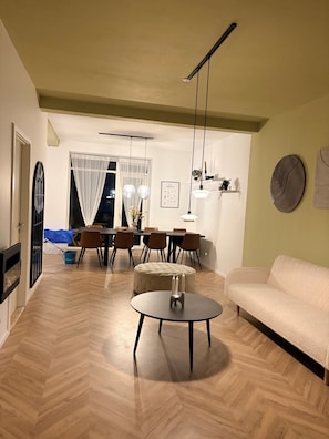En moderne stue med et rundt sofabord, en sofa, et spiseområde med stole, og et køkken i baggrunden.