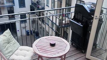 Ein Balkon mit rundem Tisch, zwei gepolsterten Stühlen, einem Grill und Blick auf angrenzende Gebäude.