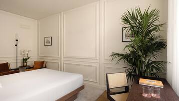 Une chambre d’hôtel avec un lit, une chaise, une table avec un livre et des lunettes, une plante en pot et un tableau au mur.