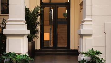 L’entrée d’un immeuble est marquée par un escalier menant à une double porte, flanquée de deux colonnes et agrémentée de plantes en pot.