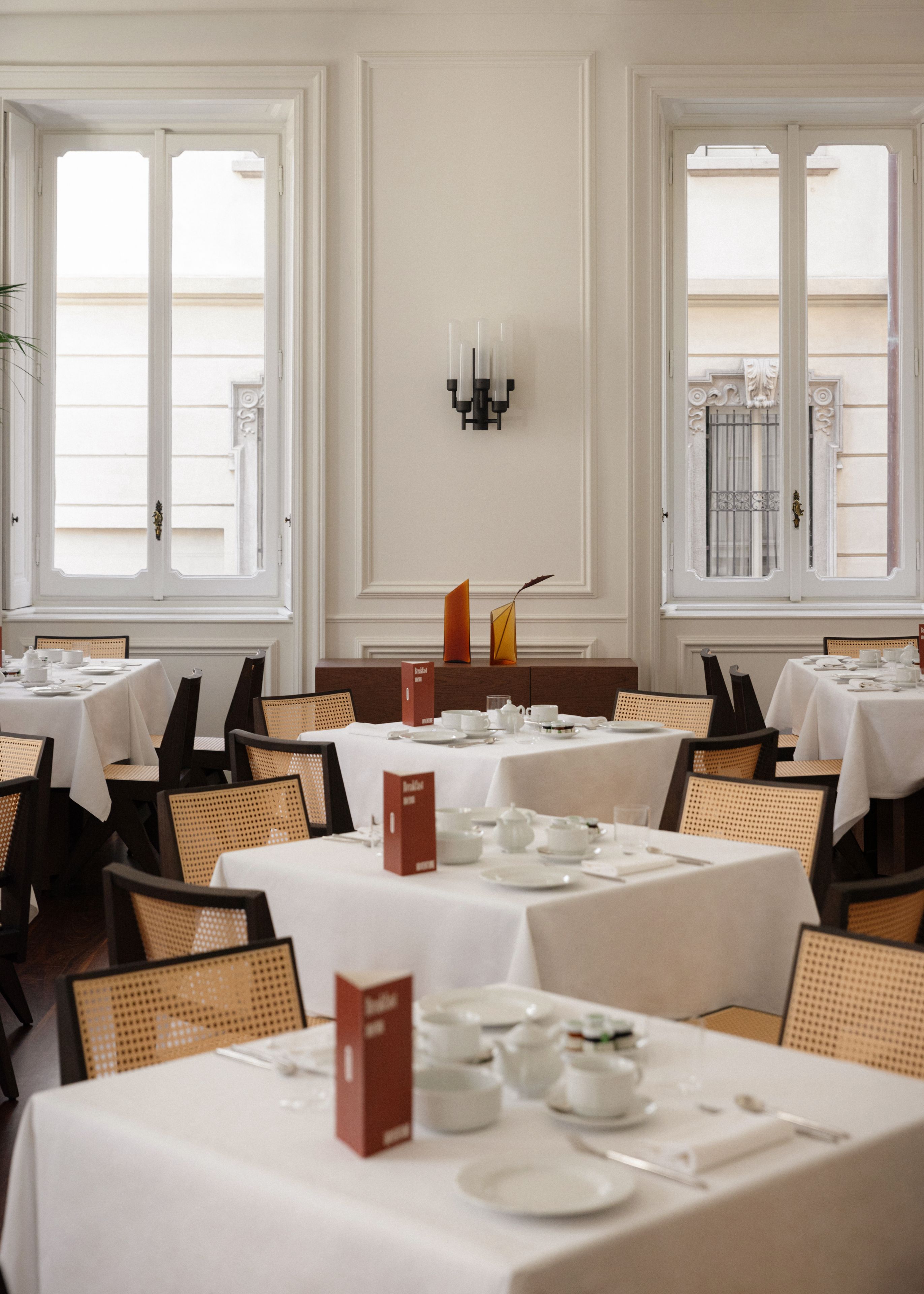Un espace repas avec plusieurs tables dressées pour le petit-déjeuner, agrémenté de nappes blanches, de chaises en osier et de différents éléments de service pour le petit-déjeuner.