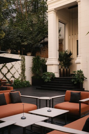 An outdoor seating area with orange sofas and tables, surrounded by greenery and a white building with columns. - Ouverture (Milan)