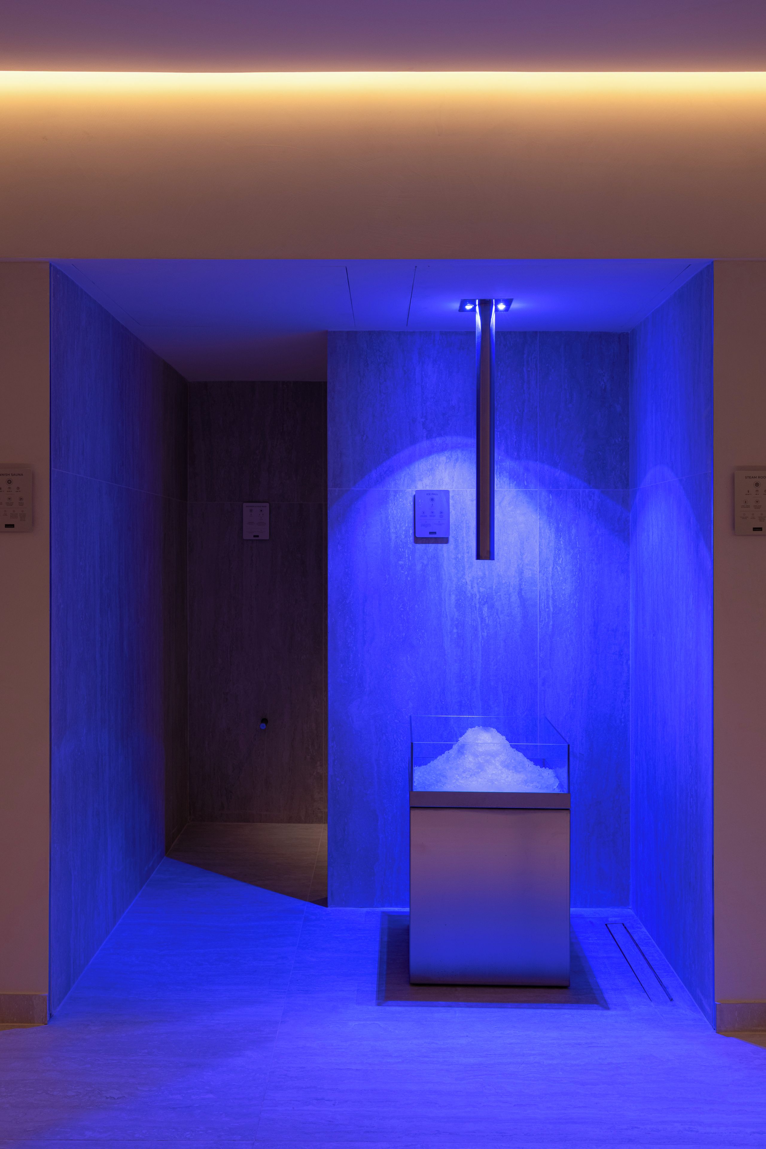 Une salle de bain moderne avec une zone de douche éclairée en bleu, un panneau de commande visible et une porte donnant sur une autre pièce.