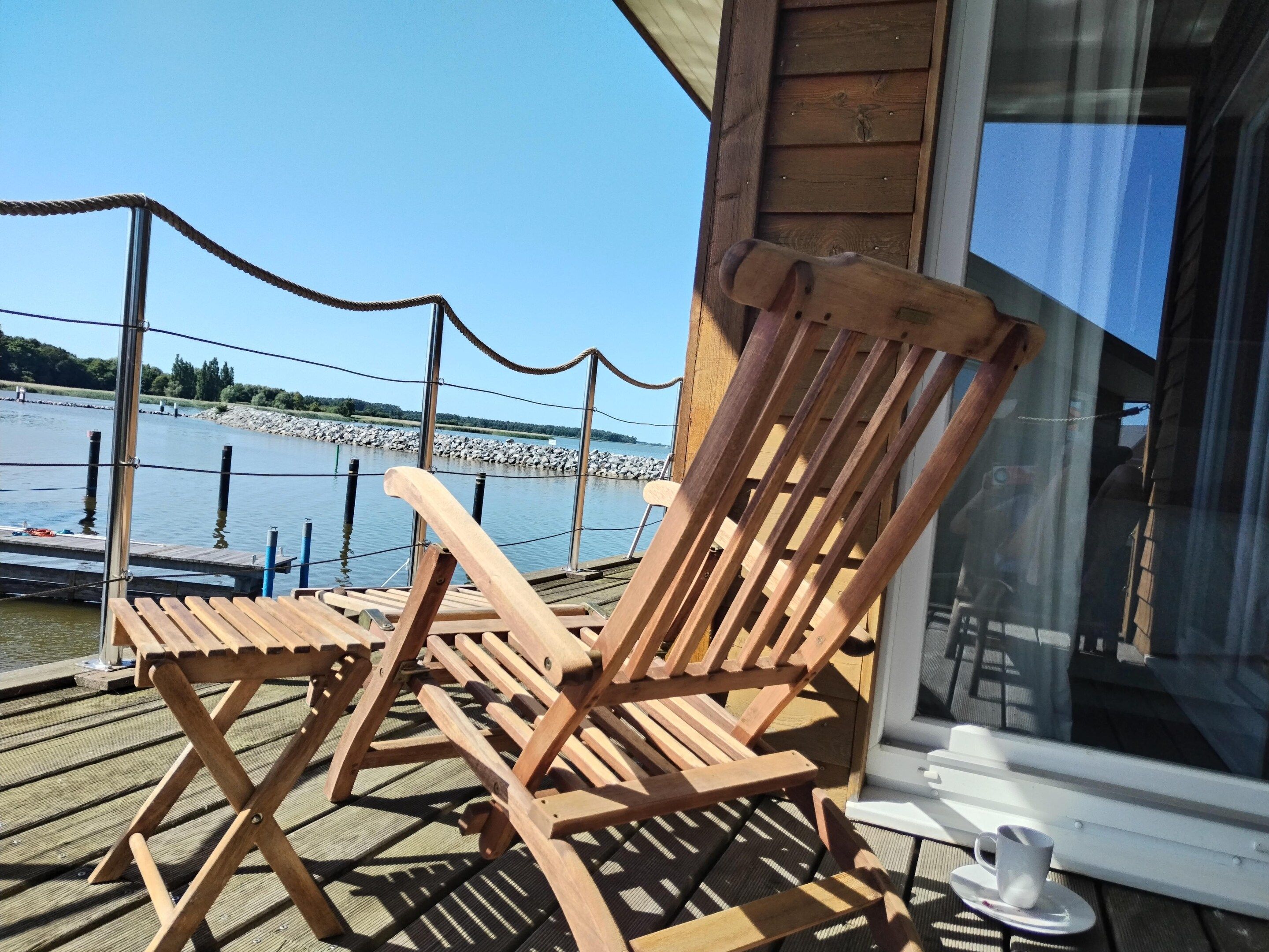 Une terrasse en bois avec deux chaises longues, une porte vitrée et une vue sur une étendue d’eau calme.