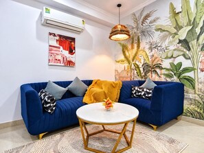 Sala de estar com um sofá azul, uma mesa redonda branca e um mural tropical na parede.