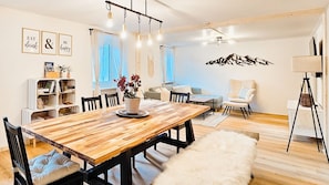 Un coin repas avec une table en bois, des chaises et un vase de fleurs. On y trouve également une bibliothèque, un canapé et une lampe.