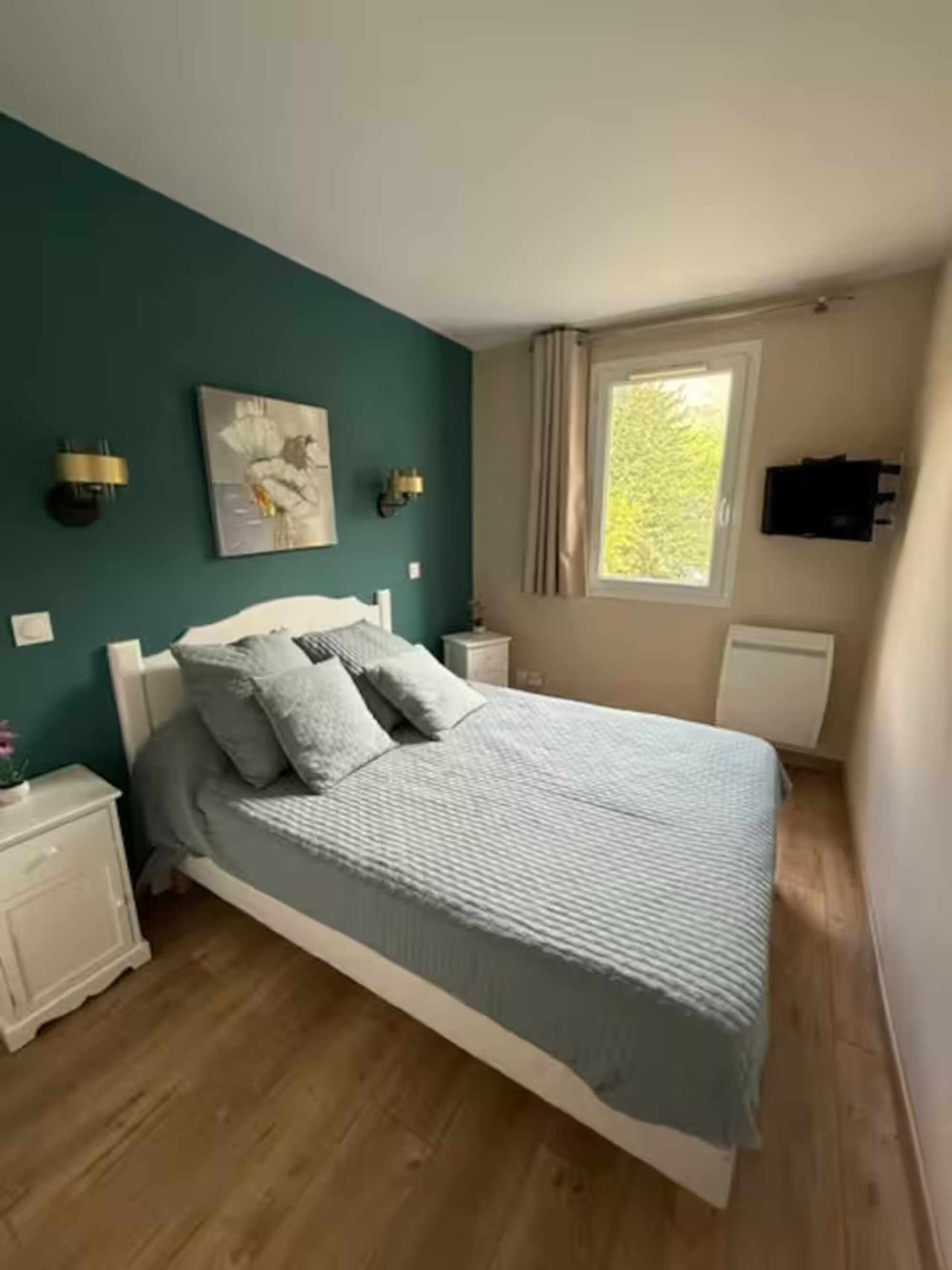 Ein Schlafzimmer mit einem Bett, einem Nachttisch, einer Wandlampe, einem Fenster mit Vorhängen und einem Bild an der Wand.