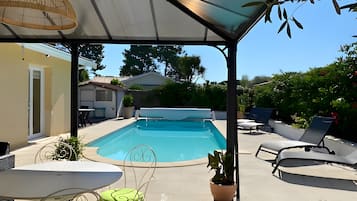 A covered outdoor area with a swimming pool, lounge chairs, and a table with chairs.