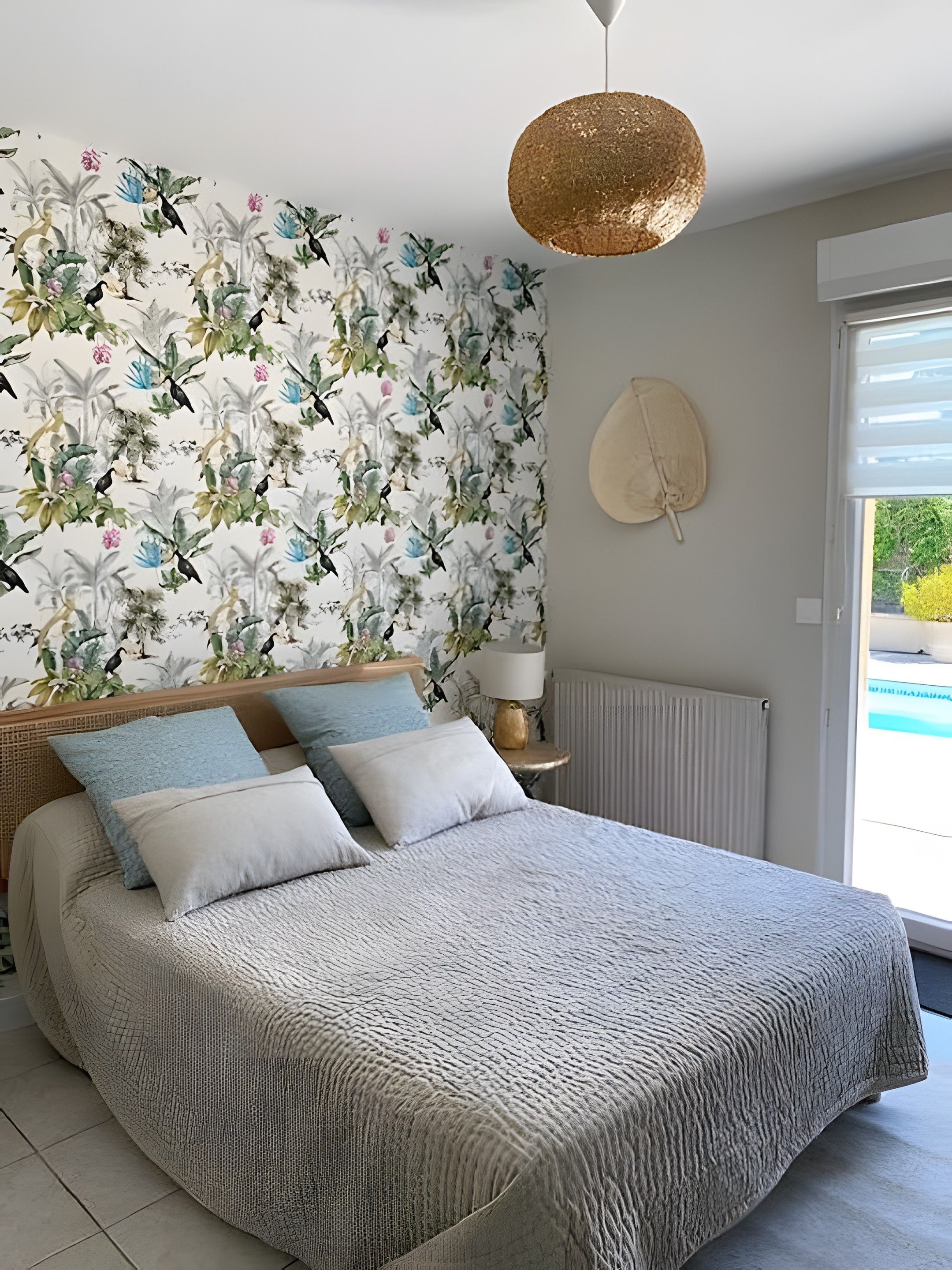 Un dormitorio con cama, pared con estampado floral, lámpara colgante y ventana con vista.