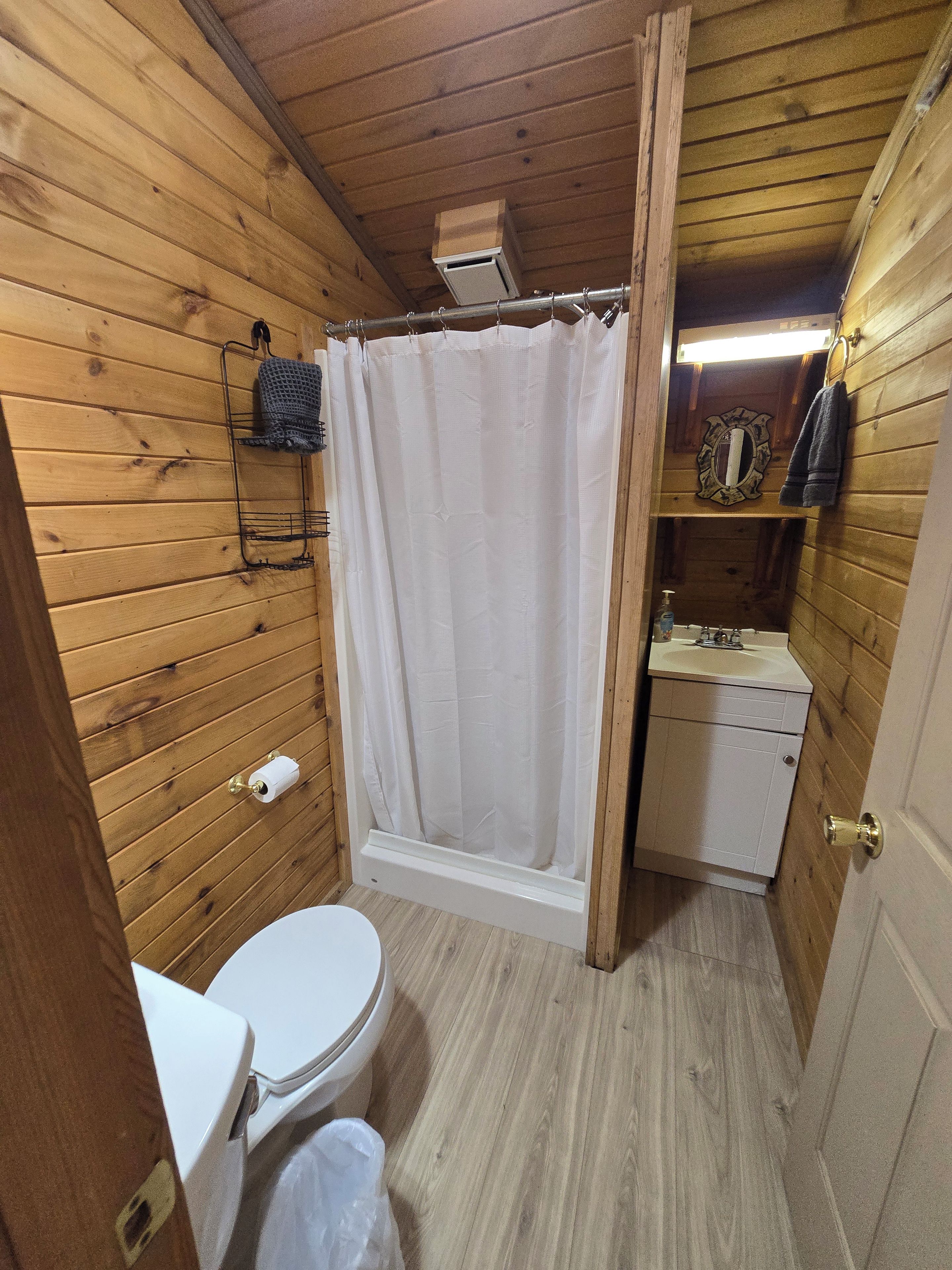 A wooden-paneled bathroom with a toilet, shower curtain, and a small sink with a mirror.