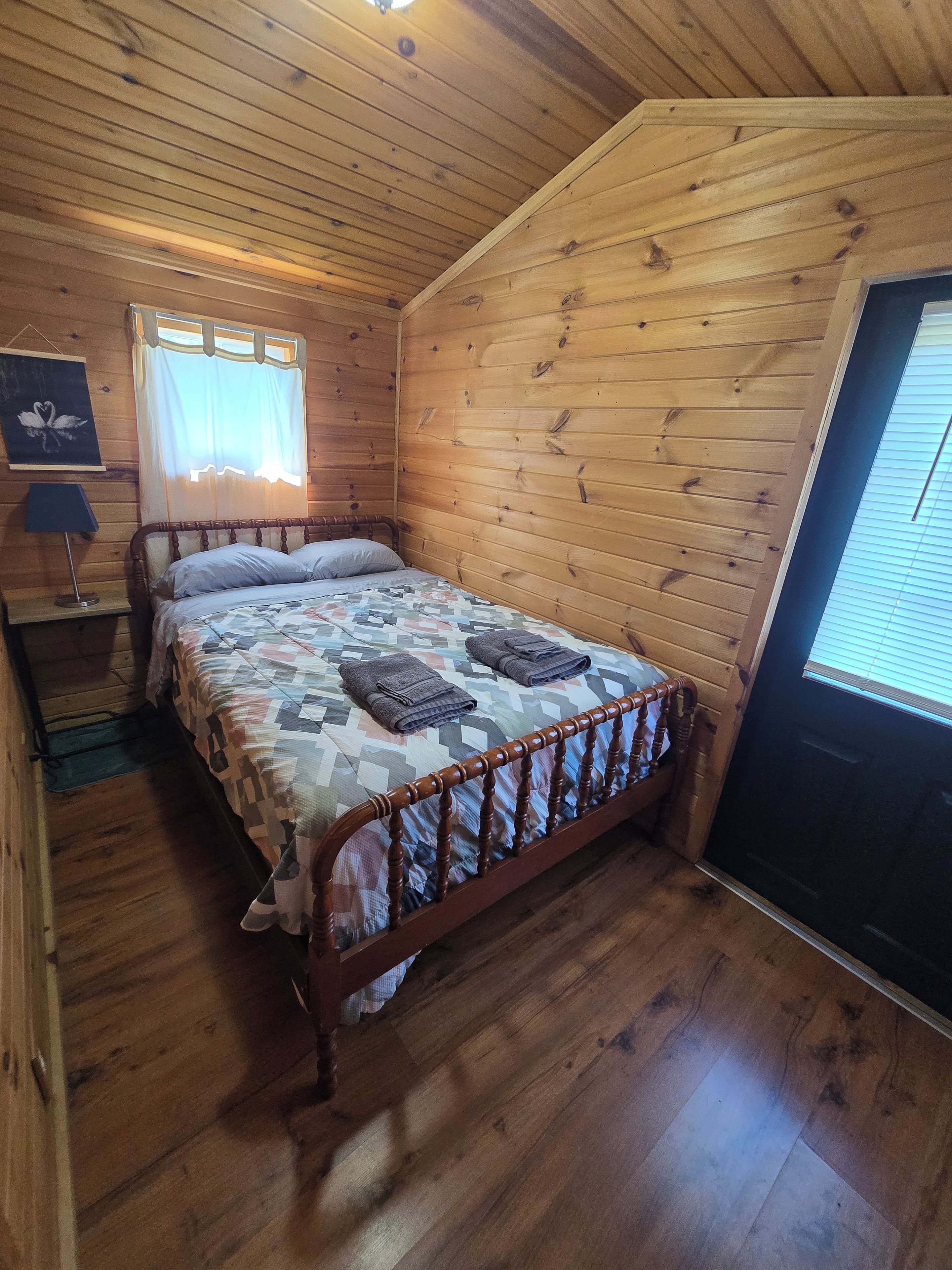A wooden-paneled room with a bed, a window with curtains, and a lamp on a bedside table.