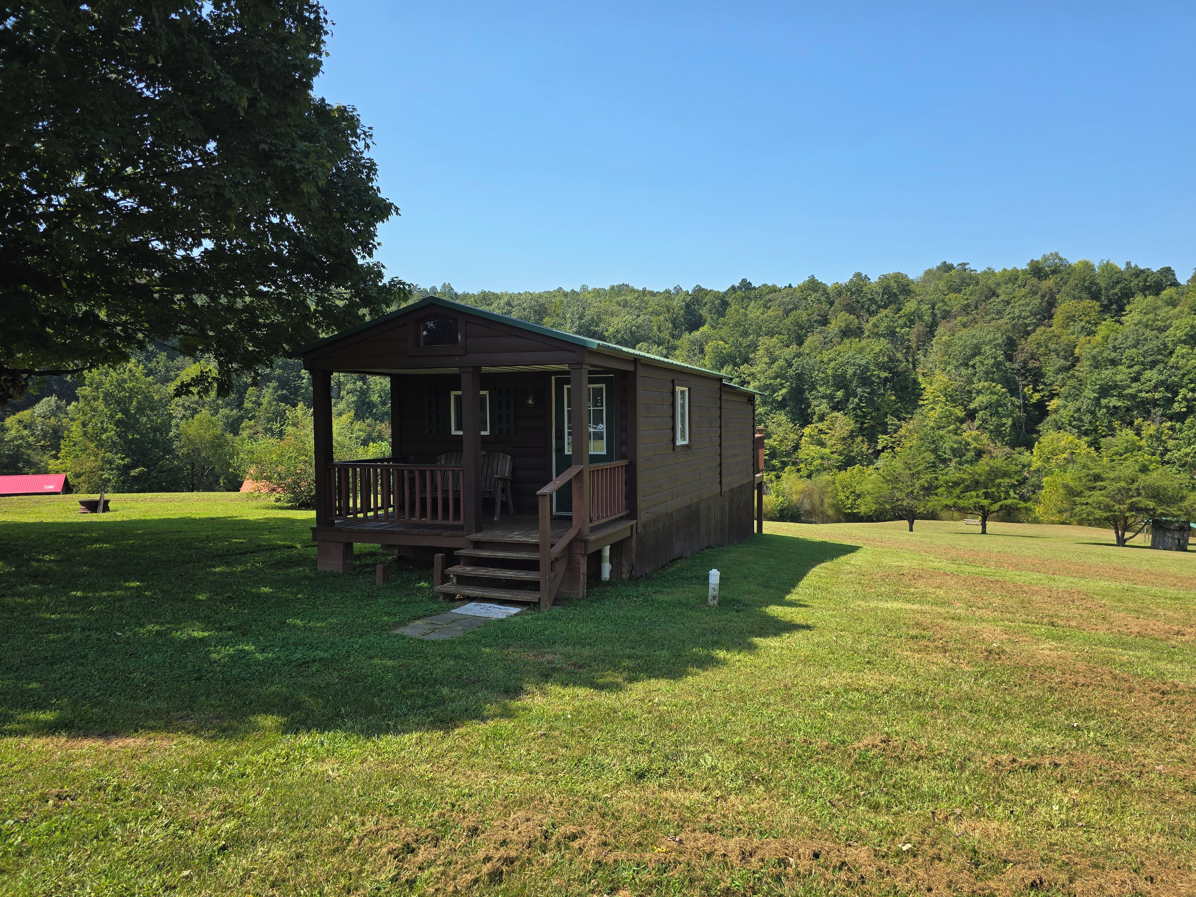 A wooden cabin with a porch, surrounded by a grassy field and dense forest.