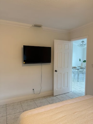 Ein Zimmer mit einem an der Wand befestigten Fernseher, einer weißen Tür und Blick in einen Essbereich mit einer Chandelier.