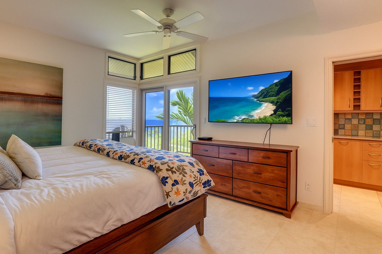 A bedroom with a bed, a TV, a ceiling fan, and a view of the ocean.