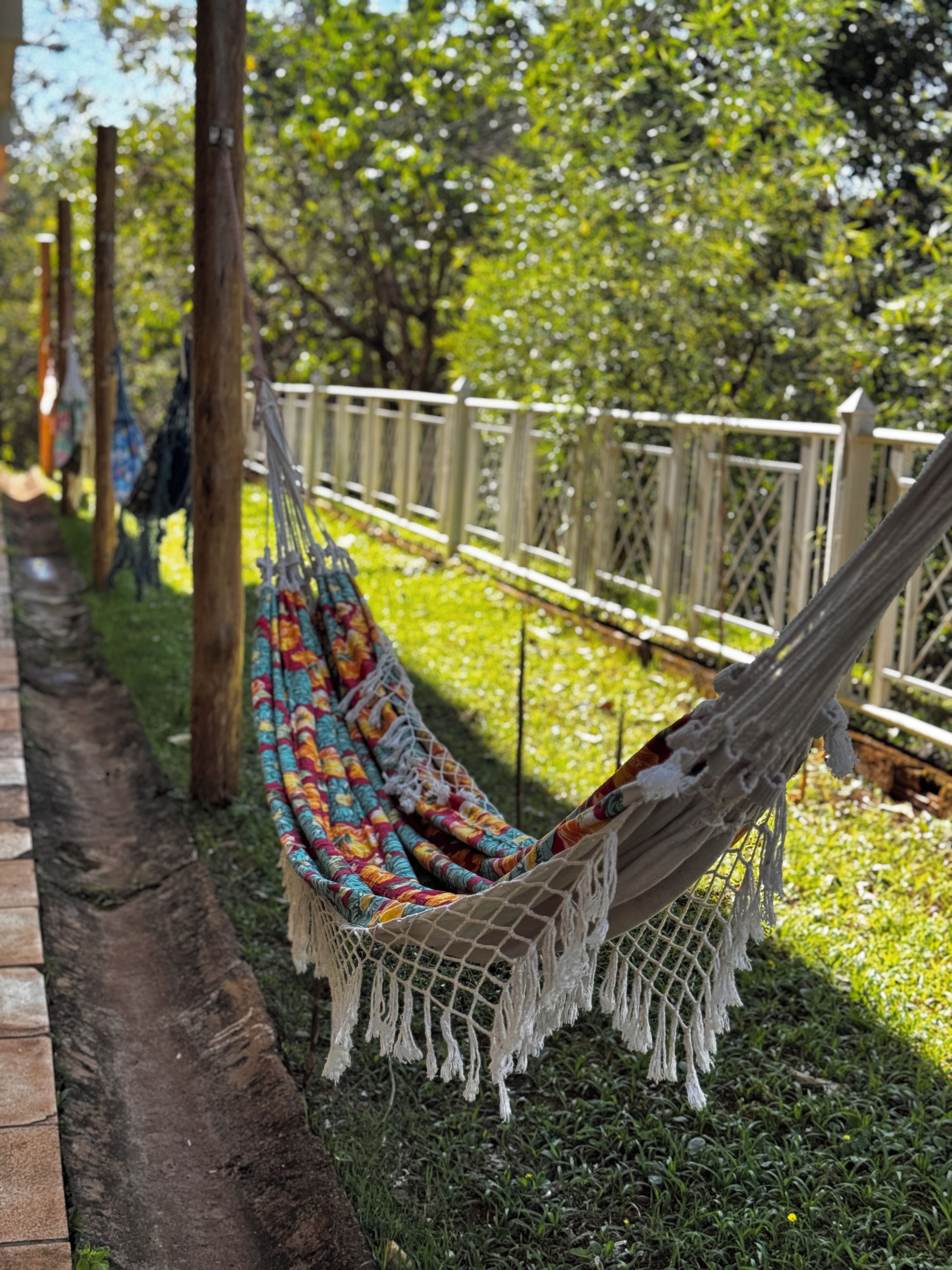 Un hamac fait d’un tissu aux motifs colorés, suspendu entre deux arbres dans une zone herbeuse, avec une clôture en bois en arrière-plan.