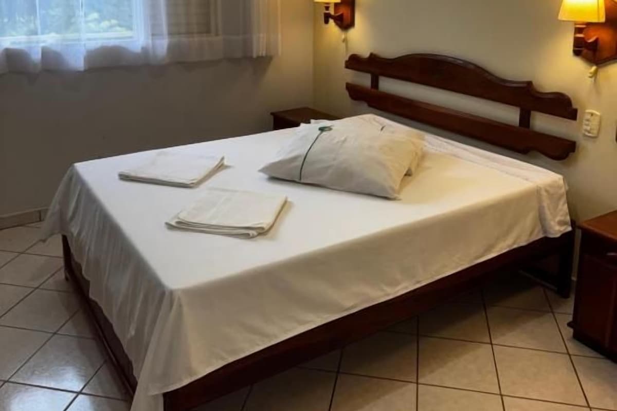 A single bed with a wooden headboard, two bedside lamps, a window with sheer curtains, and a tiled floor.