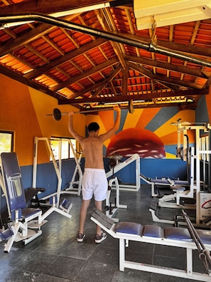 A gym with various exercise equipment, a person lifting weights, and a colorful wall design. - Pousada Paraíso dos Sonhos (Corumbá de Goiás)