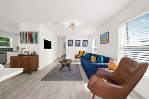 Un salón moderno con un sofá, una mesa de centro y un sillón de cuero marrón. Se observa una zona de cocina con un armario de madera y una mesa de comedor.