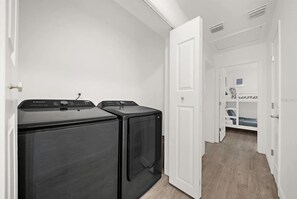 Een wasserette met twee zwarte wasmachines en drogers, een witte deur, en een gang naar een slaapkamer met stapelbedden.