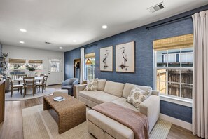 A modern living room with a blue accent wall, a beige sofa, a dining area with a table and chairs, and artwork on the wall. - Sandy Shores (Galveston)