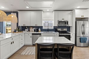 A modern kitchen with white cabinetry, a stainless steel refrigerator, a built-in oven, and a microwave. There is a dining area with a white table and two chairs. - Sandy Shores (Galveston)