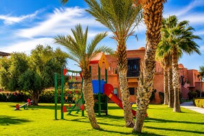 Ein Spielplatz mit Klettergerüsten und Palmen in einer angelegten Grünfläche, im Hintergrund ein Gebäude.