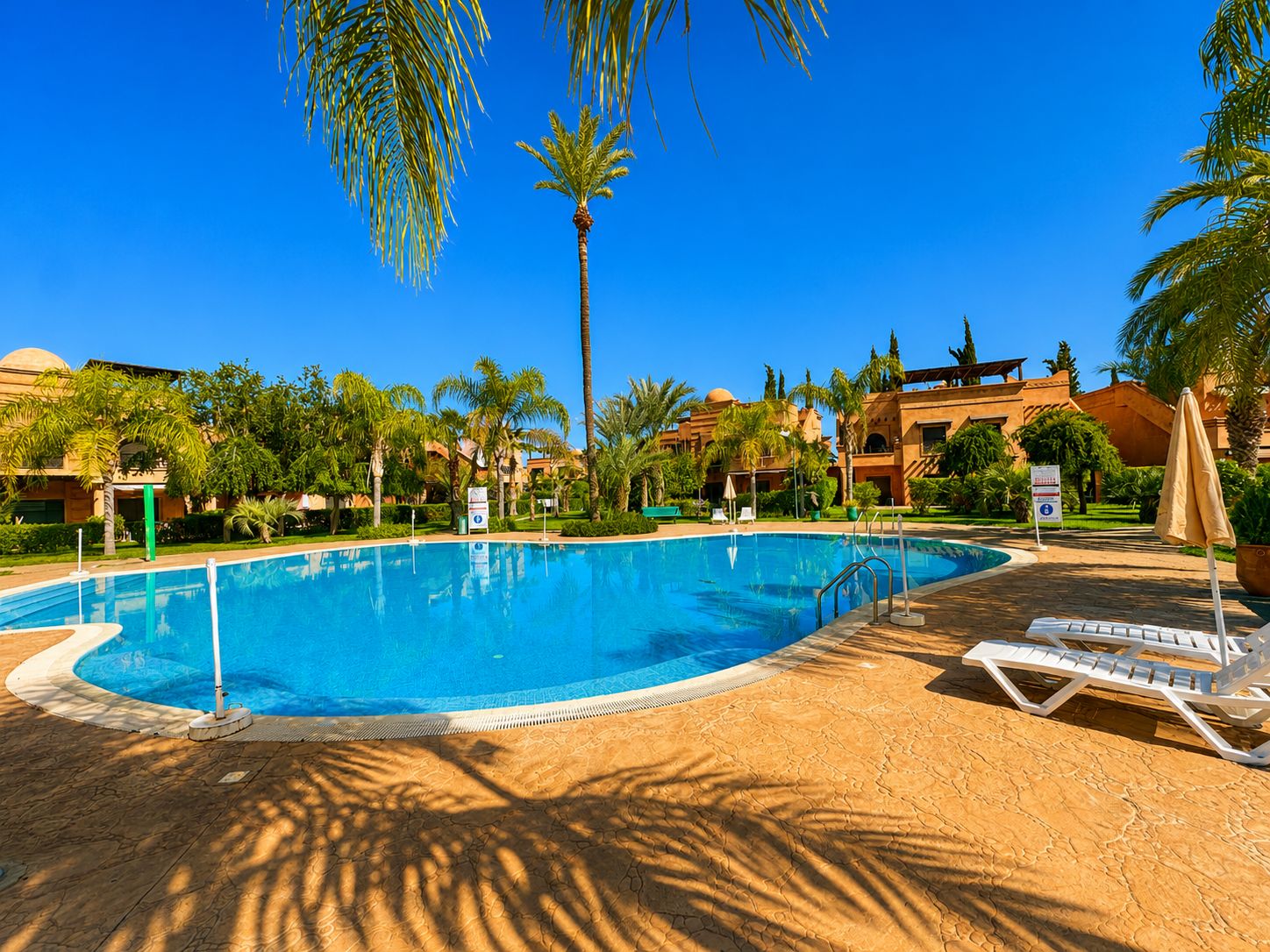 A swimming pool with lounge chairs and palm trees, surrounded by buildings with a desert landscape.