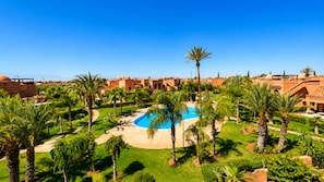 Un complex turístic amb una piscina central envoltada de palmeres i un cel blau clar.