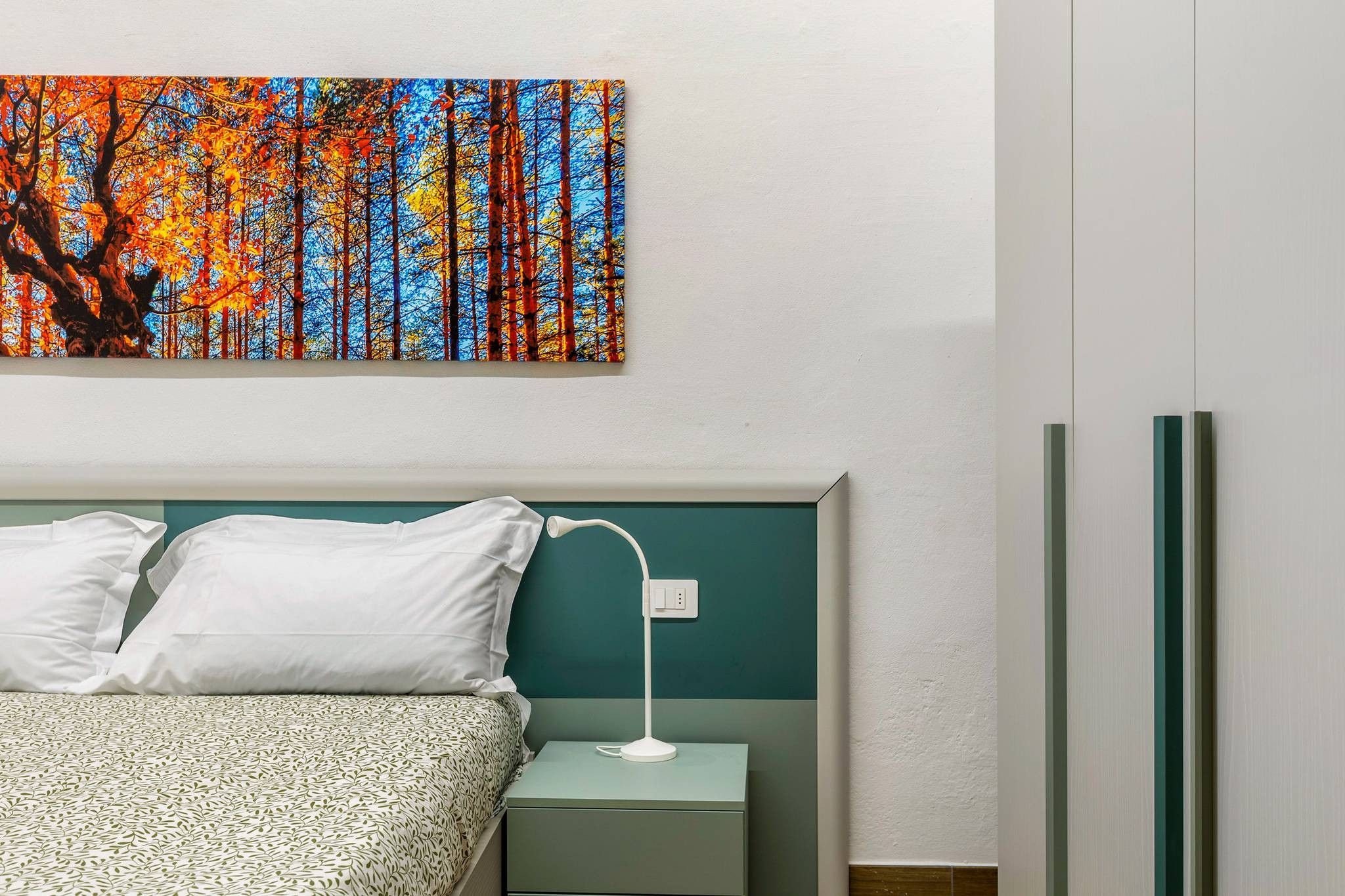 En snyggt bäddad säng med vita kuddar och ett mönstrat överkast, ett sängbord med en lampa och en färgstark, abstrakt tavla på väggen.