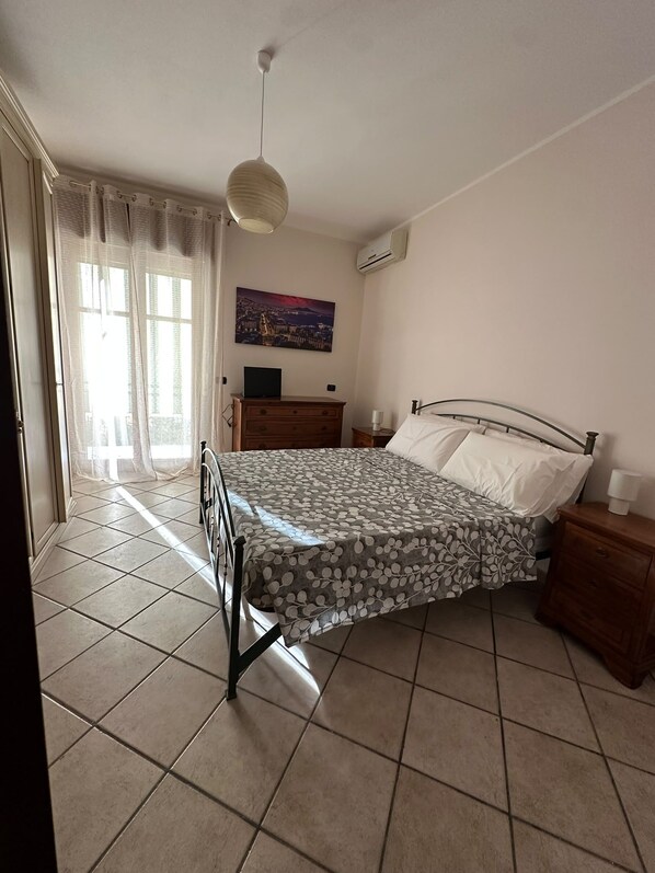 Een slaapkamer met een bed, een nachtkastje, een kapstok, een raam met gordijnen en een airconditioning.