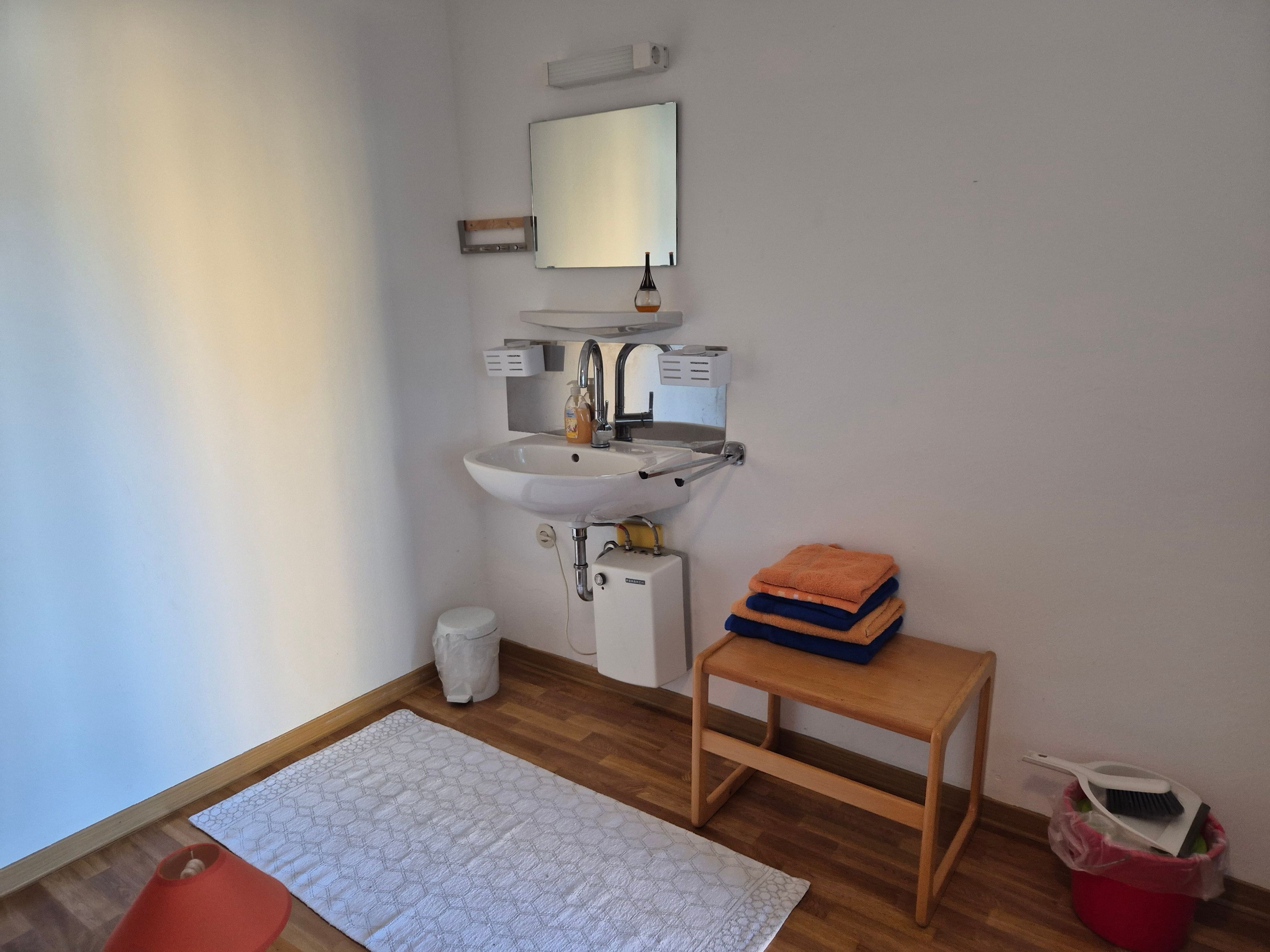 Ein kleines Badezimmer mit Waschbecken, Spiegel und einem hölzernen Hocker mit Handtüchern.